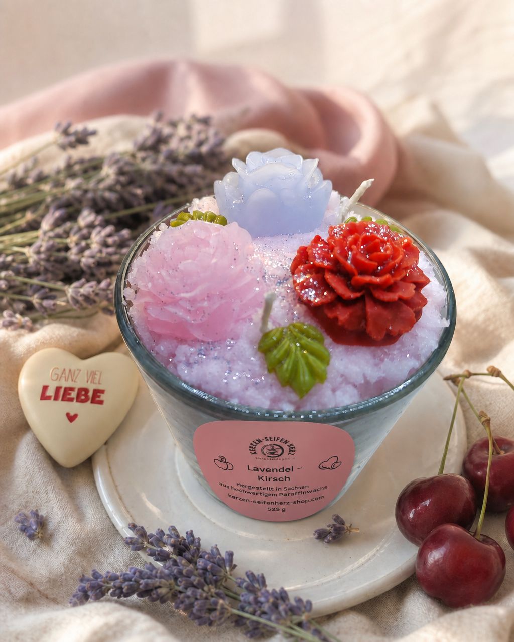 Lavendel-Kirsch Duftkerze auf heller Steinplatte mit rosa Tuch, Lavendel und Kirschen