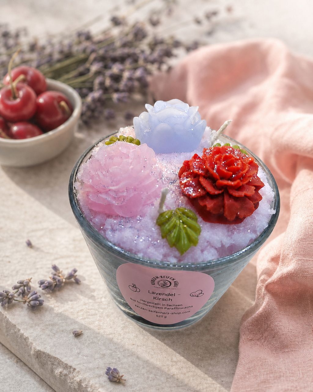 Lavendel-Kirsch Duftkerze auf heller Steinplatte mit rosa Tuch, Lavendel und Kirschen