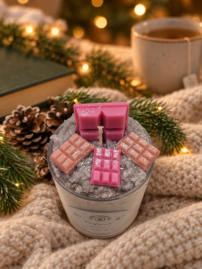 Tiramisu-Weihnachtskerze im Glas mit rosa Schokoladendekoration auf Strickdecke neben Tasse und Weihnachtslichtern